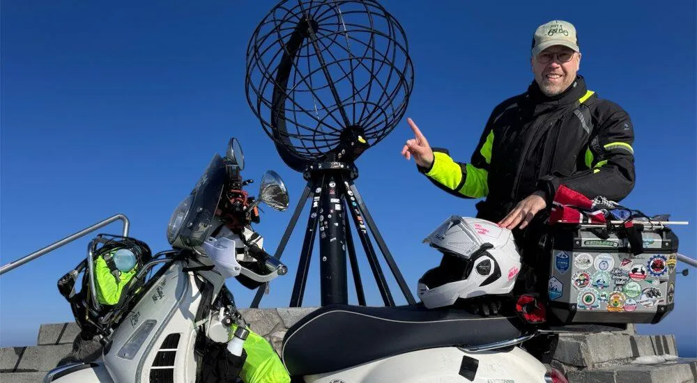 Olaf Just mit seiner beladenen weißen Vespa GTS 300 stehend vor dem Globe am Nordkapp bei strahlend sonnigem Wetter